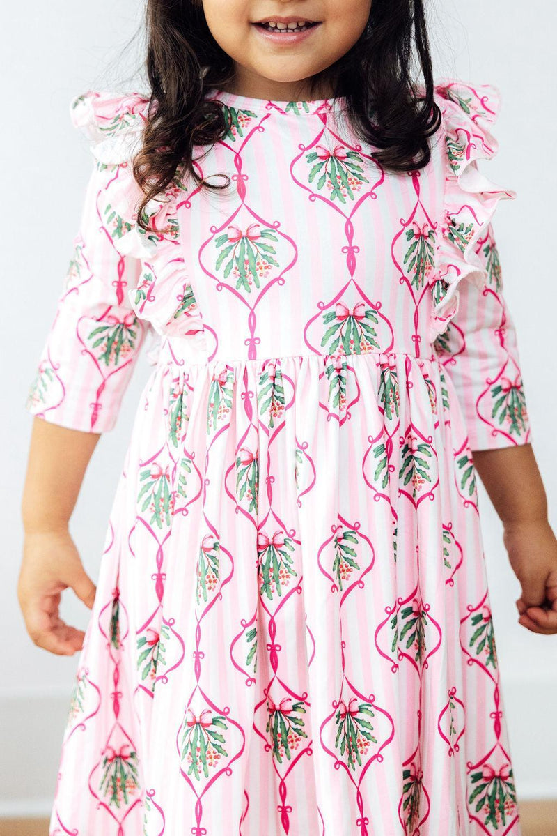 A joyful child wearing the Merry Mistletoe 3/4 Sleeve Ruffle Twirl Dress, showcasing a pink mistletoe pattern with ruffles, perfect for holiday festivities.