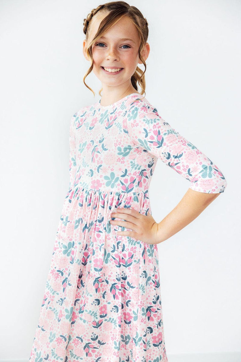 Smiling girl with hands on her hips wearing the Softly Blooming 3/4 Sleeve Pocket Twirl Dress by Mila & Rose®, displaying the vibrant floral print and charming silhouette.