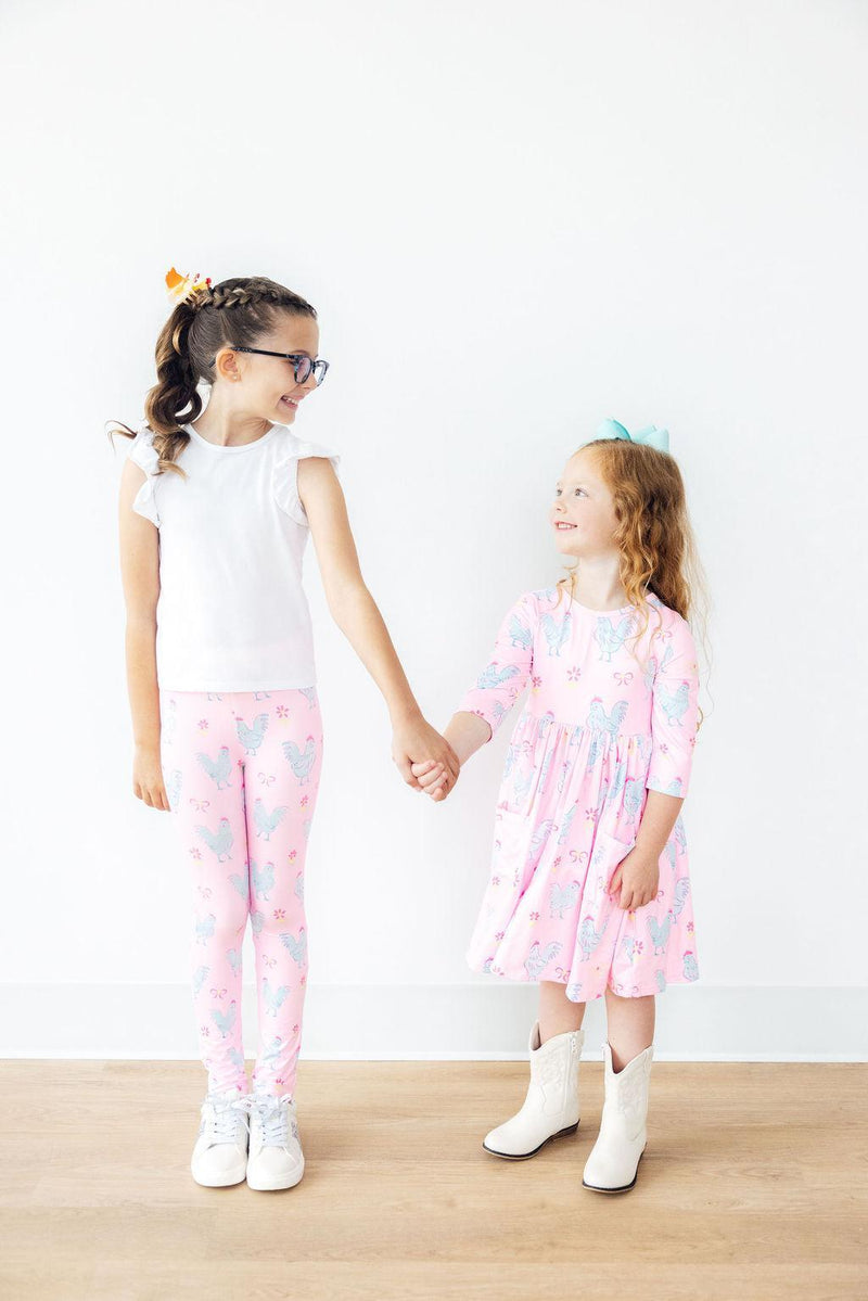 Two girls holding hands, one in the Little Miss Chickie 3/4 Sleeve Pocket Twirl Dress and the other in playful leggings, demonstrating friendship and fun in stylish outfits.
