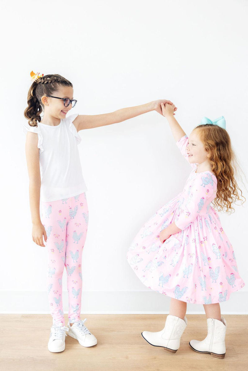 Two girls enjoying a playful moment, one wearing the Little Miss Chickie 3/4 Sleeve Pocket Twirl Dress and the other in cute leggings, highlighting the dress's versatility for fun occasions.