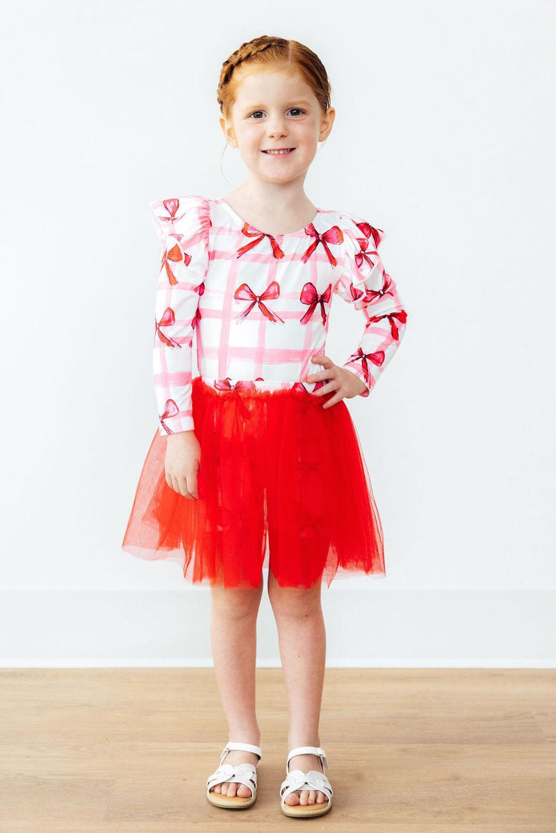 A young girl wearing Bow La La Tutu Shorts by Mila & Rose®, featuring a playful red skirt and a pink top with bows, proudly posing with her hand on her hip, showcasing her joyful style.