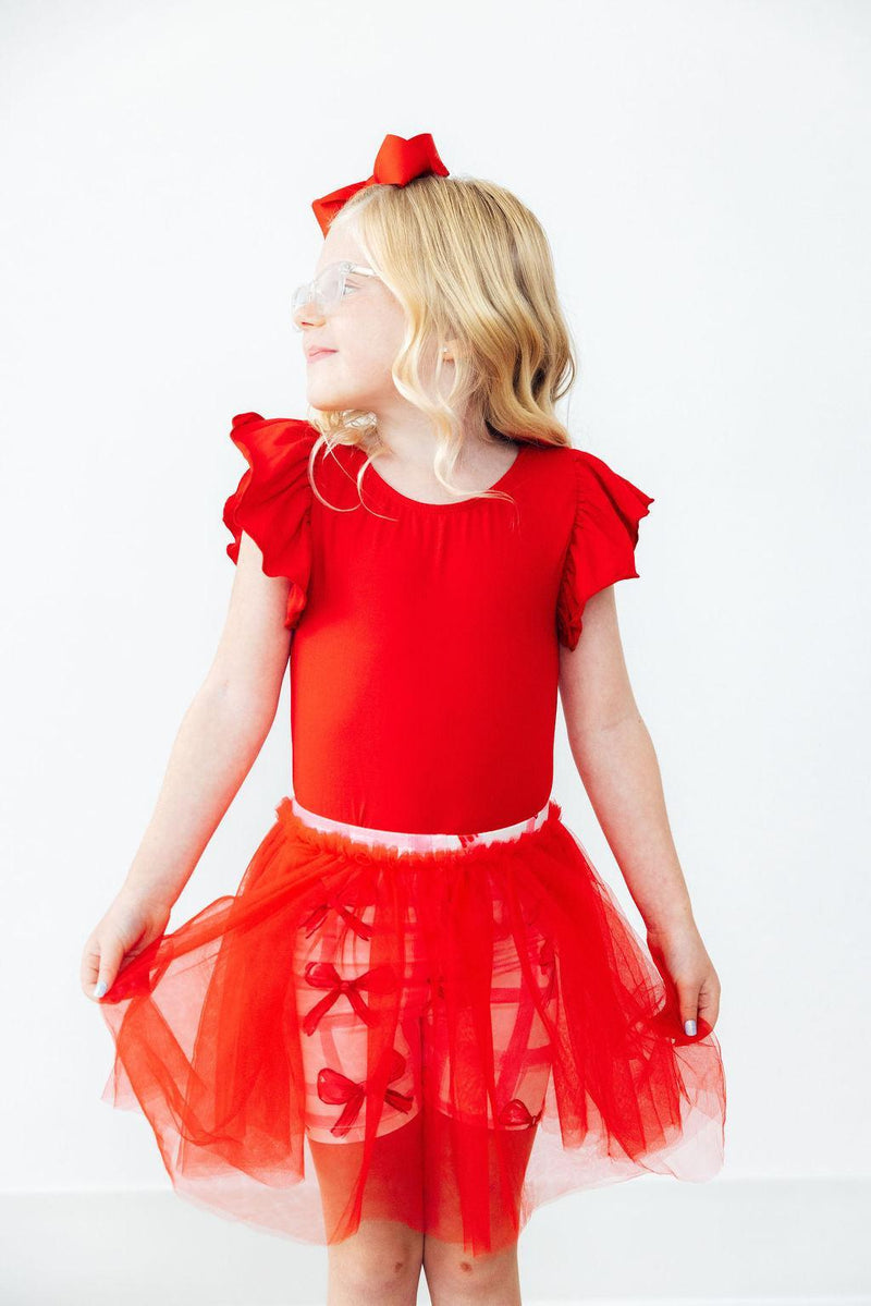 A smiling girl wearing the Bow La La Tutu Shorts and a red top, highlighting the cheerful design of the tutu and its vibrant color, ideal for playful occasions.