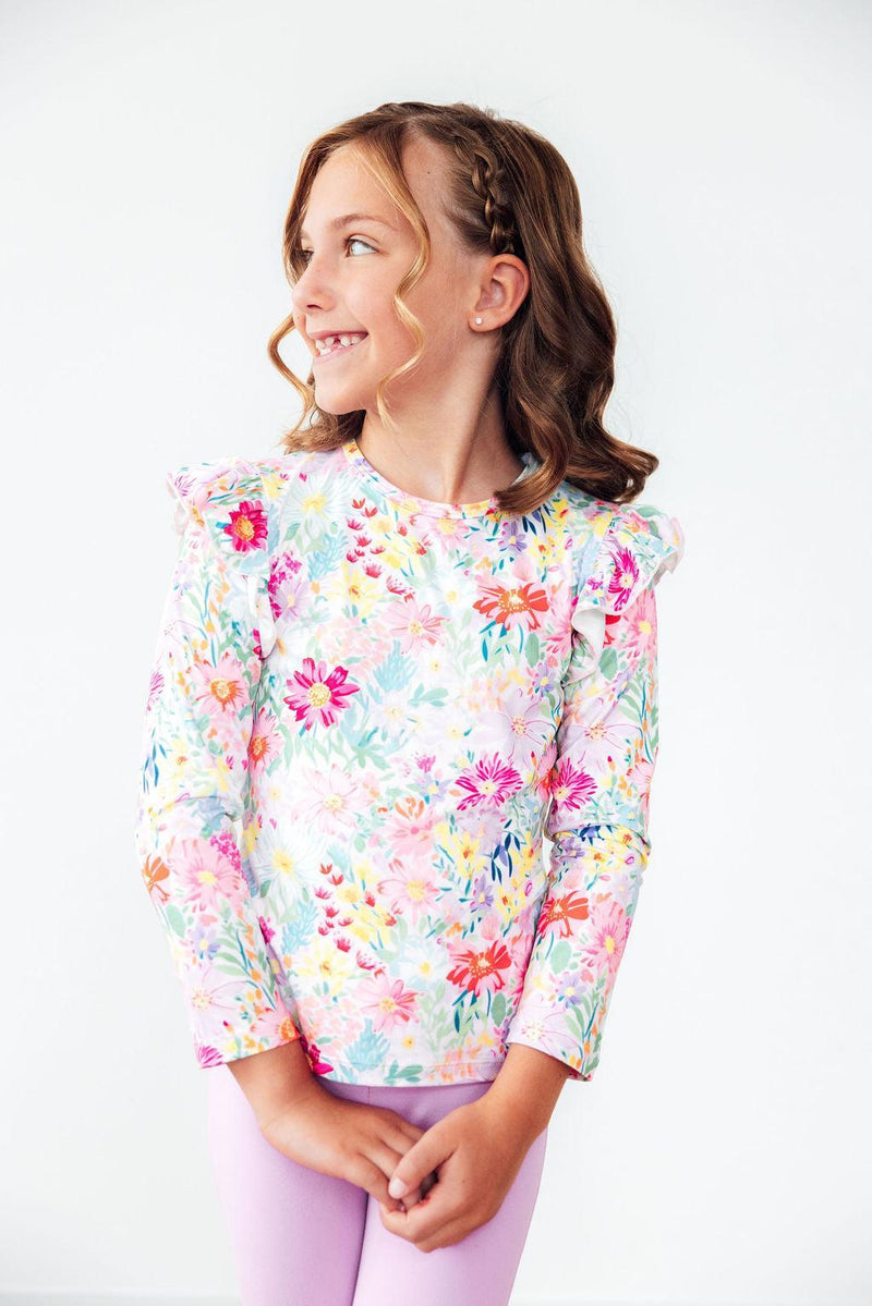 Smiling girl wearing the Field of Flowers L/S Ruffle Tee from Mila & Rose, showcasing vibrant floral prints and ruffled sleeves, perfect for twirling and playtime.