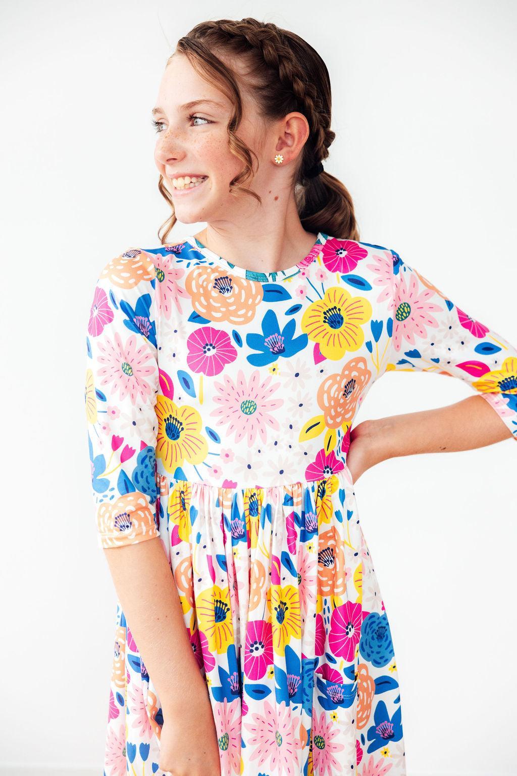 A cheerful girl wearing the 'Sunshine Floral' 3/4 Sleeve Pocket Floral Twirl Dress, showcasing its vibrant colors and playful design, embodying the essence of a floral twirl dress.