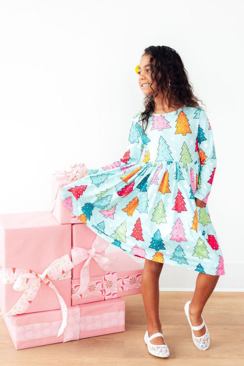 A cheerful girl wearing the Oh Christmas Tree L/S Pocket Twirl Dress, surrounded by pink gift boxes, showcasing the dress's festive tree print and twirl-worthy design. Perfect for the holiday season!