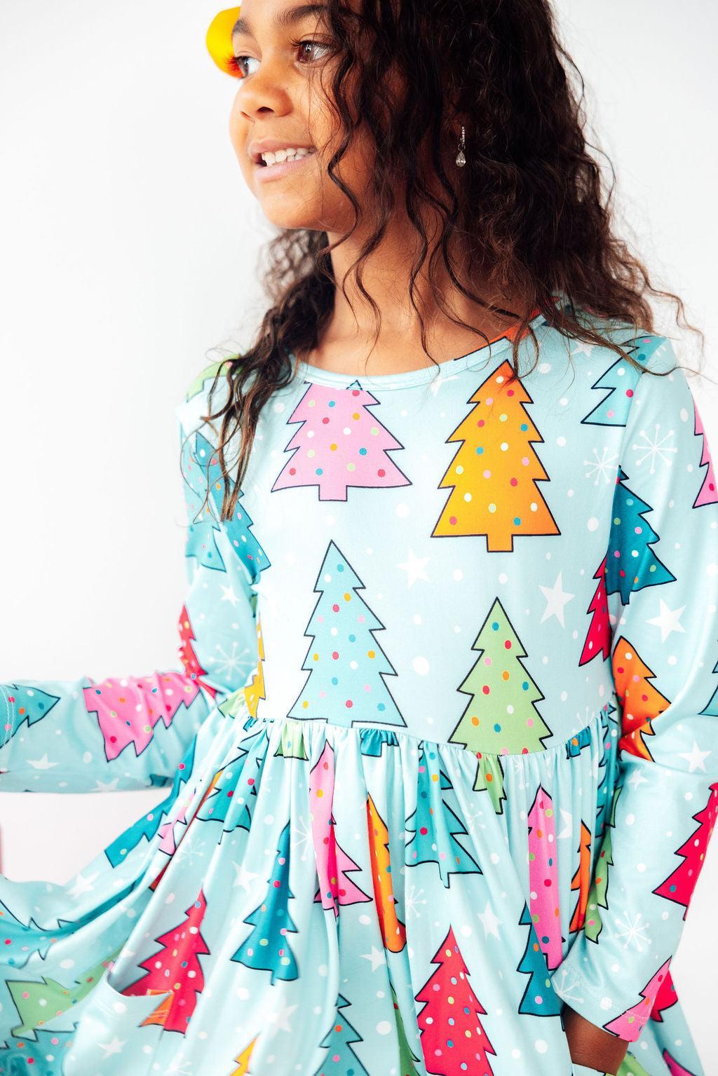 A joyful child wearing the 'Oh Christmas Tree L/S Pocket Twirl Dress' featuring colorful Christmas tree patterns against a light blue background, perfect for festive fun and twirling adventures.