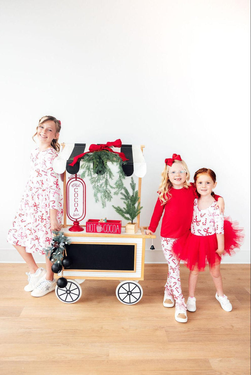 Three cheerful girls in festive attire, showcasing the Candy Cane Lane L/S Pocket Twirl Dress alongside other holiday-themed outfits, with a hot cocoa cart in the background.