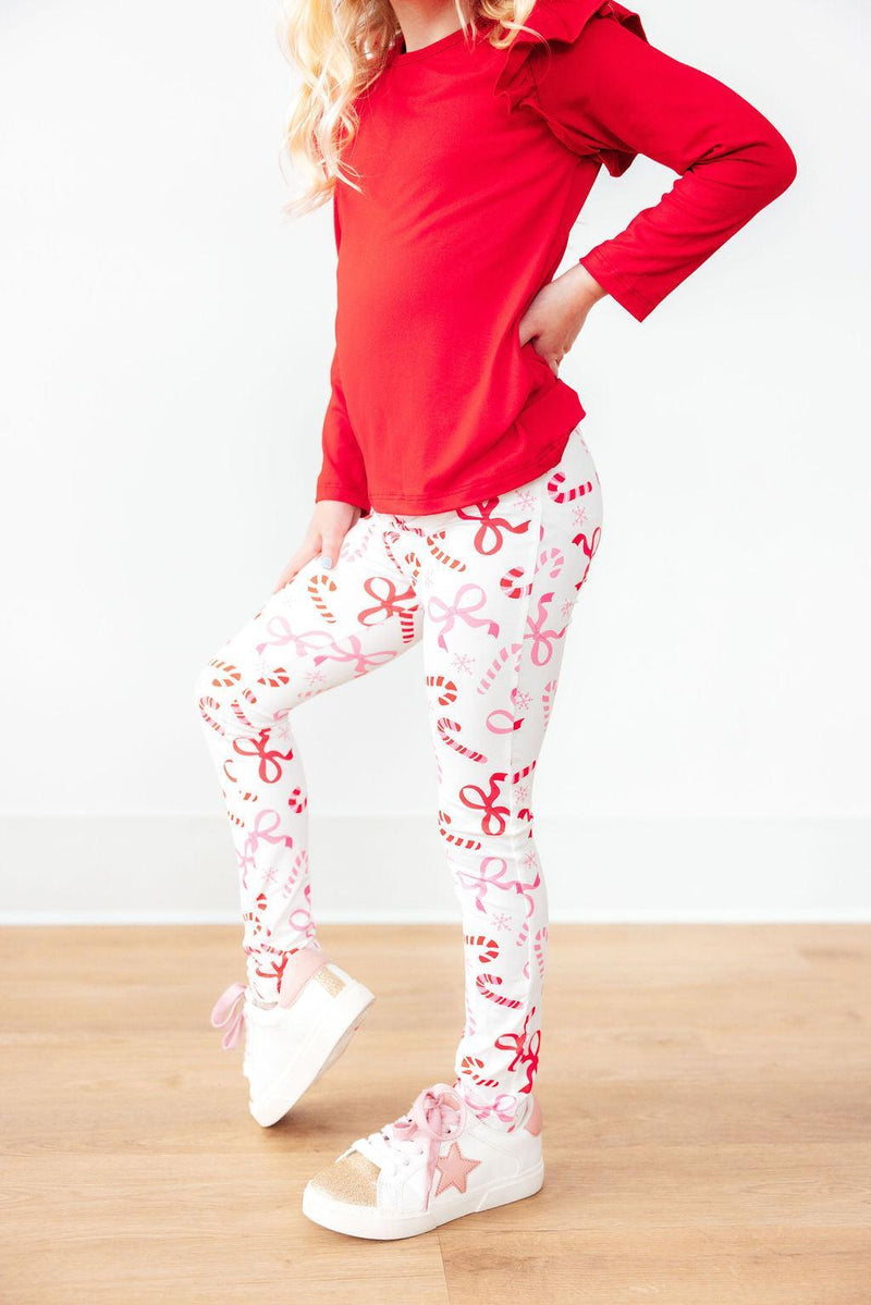 A young girl modeling Candy Cane Lane Leggings from Mila & Rose ®, featuring a festive candy cane print, paired with a red long-sleeve top and stylish white sneakers.
