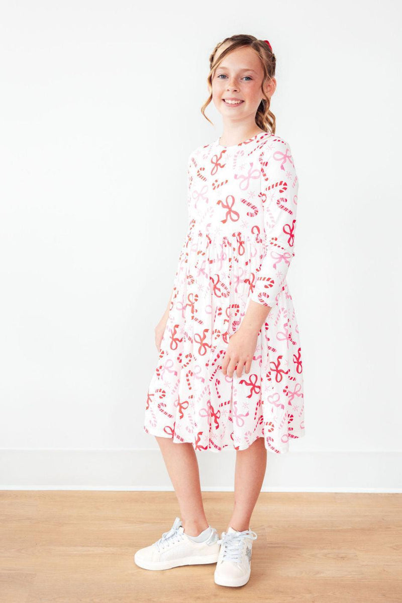 A happy girl modeling the Candy Cane Lane L/S Pocket Twirl Dress, featuring a full skirt and cute candy-themed print, perfect for twirling and holiday fun.