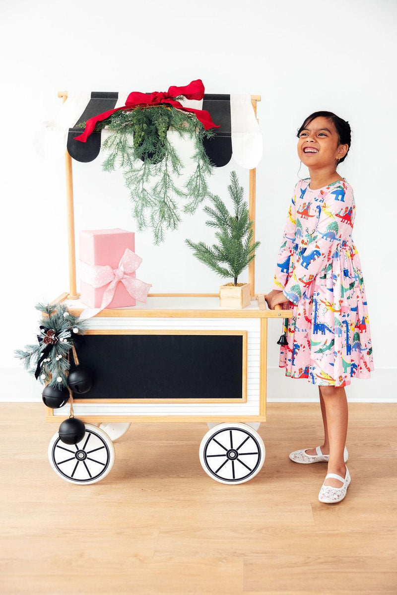 A joyful girl in an Adorable Jingle Saurus Long Sleeve Dress for Girls, playing beside a festive cart with holiday decorations, emphasizing fun and style in a long sleeve dress for girls.