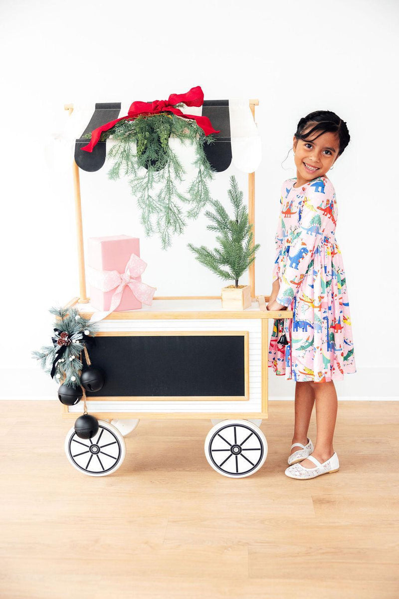 Another view of a happy girl wearing the Adorable Jingle Saurus Long Sleeve Dress for Girls, standing next to a colorful cart adorned with gifts and greenery, perfect for any occasion in a long sleeve dress for girls.