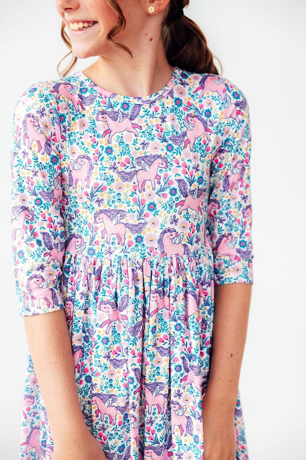 A girl smiling while wearing the Head in the Clouds 3/4 Sleeve Pocket Twirl Dress for Girls. This colorful twirl dress for girls features a whimsical unicorn and floral print, perfect for school or play.