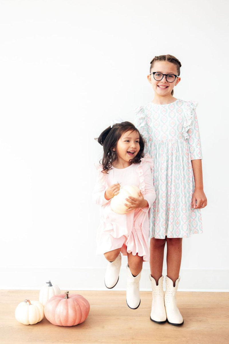 Two joyful girls, one in a Petal Pink L/S Ruffle Twirl Dress by Mila & Rose® and the other in a patterned dress, jumping with pumpkins, showcasing playful style and seasonal fun.