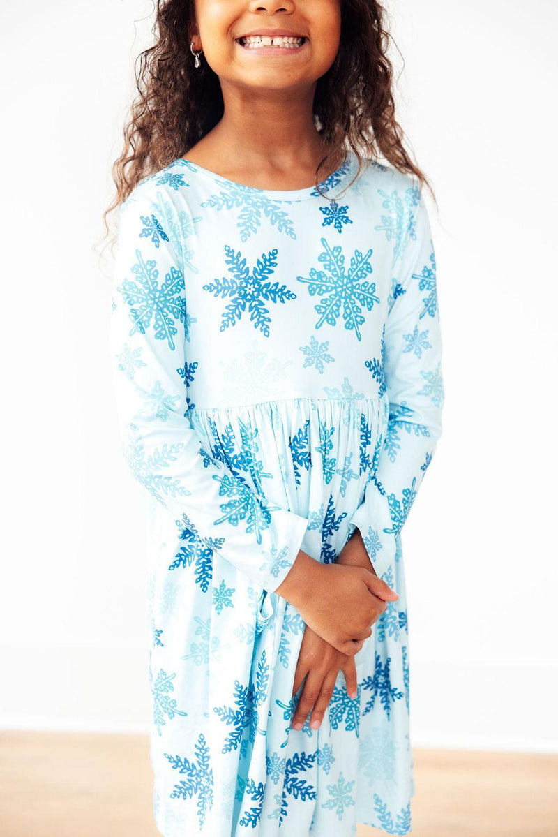 A close-up of a happy child in the Winter Wonderland L/S Pocket Twirl Dress by Mila & Rose, featuring a charming blue snowflake pattern and a playful pose.