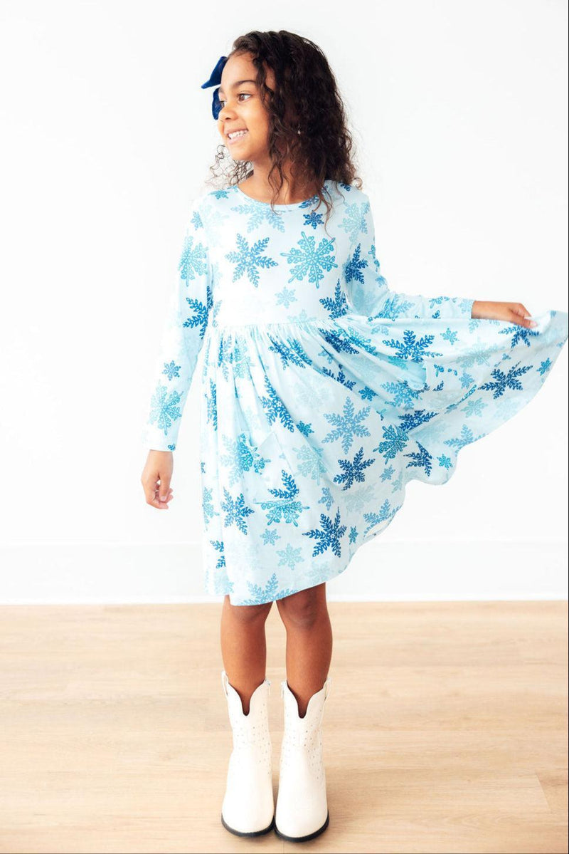 A smiling girl spinning in the Winter Wonderland L/S Pocket Twirl Dress by Mila & Rose, with a lively blue snowflake design, complemented by stylish white boots.