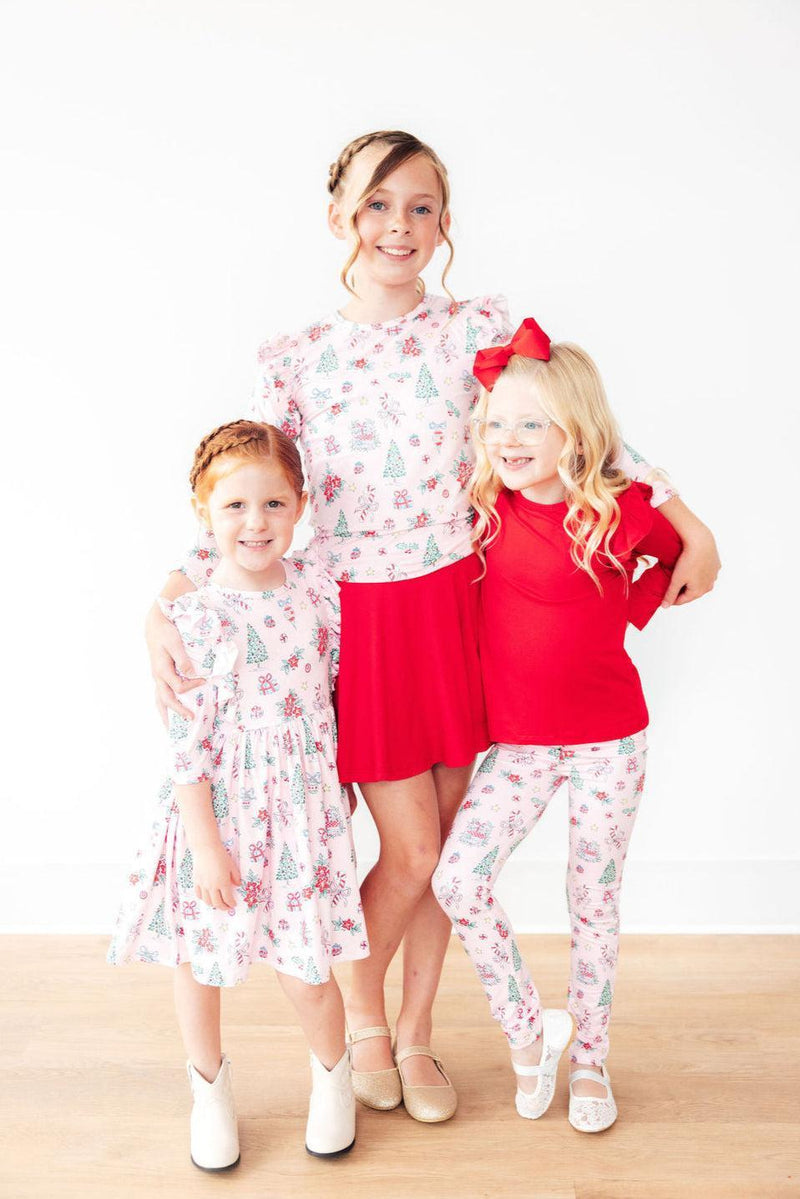 Three girls modeling pink Christmas leggings, smiling together in a playful pose, wearing coordinated festive tops. The pink Christmas leggings feature cheerful holiday patterns.