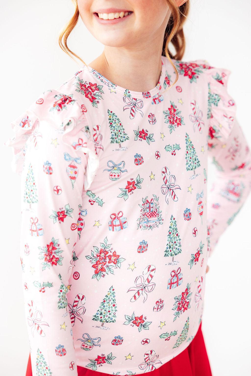 Smiling girl wearing the Pink Christmas Long Sleeve Ruffle Tee, featuring festive print with trees, gifts, and candy canes, styled with a red skirt.