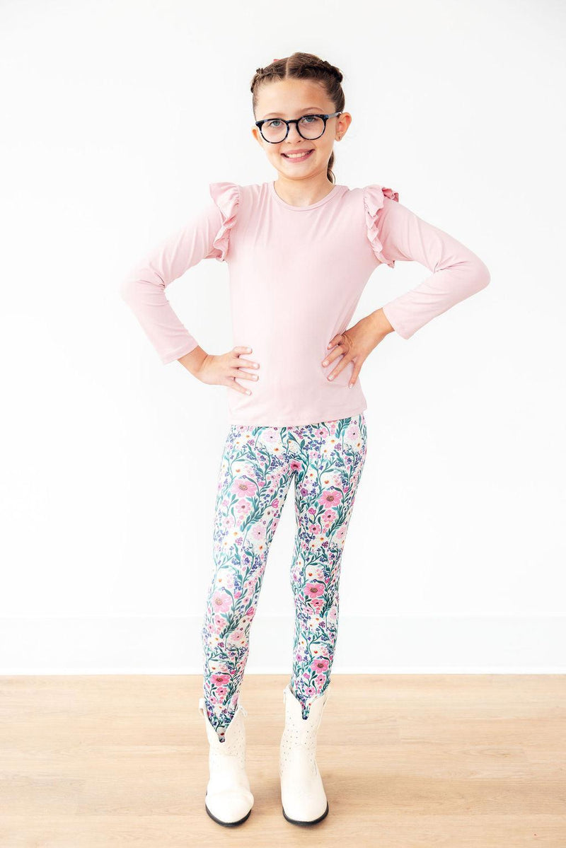 Vivid Garden Leggings showcased by a smiling girl wearing a pink ruffle top and white boots, displaying a vibrant floral print for a fun, fashionable look.