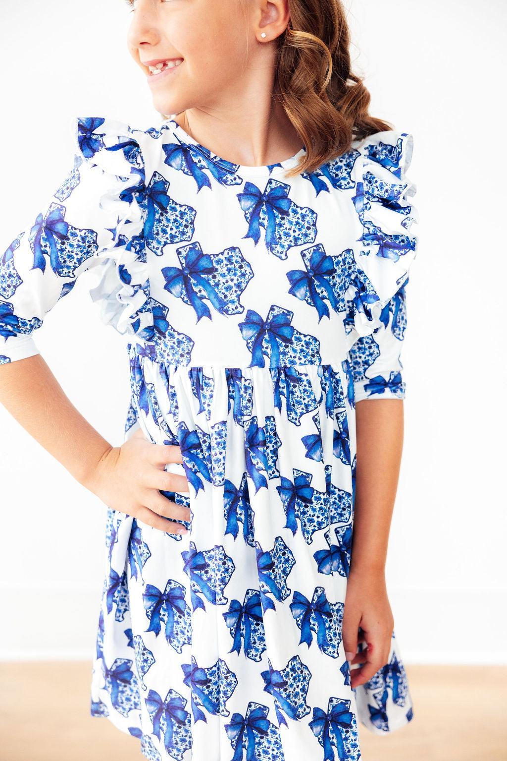 A cheerful girl modeling the Lone Star 3/4 Sleeve Ruffle Twirl Dress with a playful blue bow pattern, showcasing its stylish ruffles and comfortable fit.