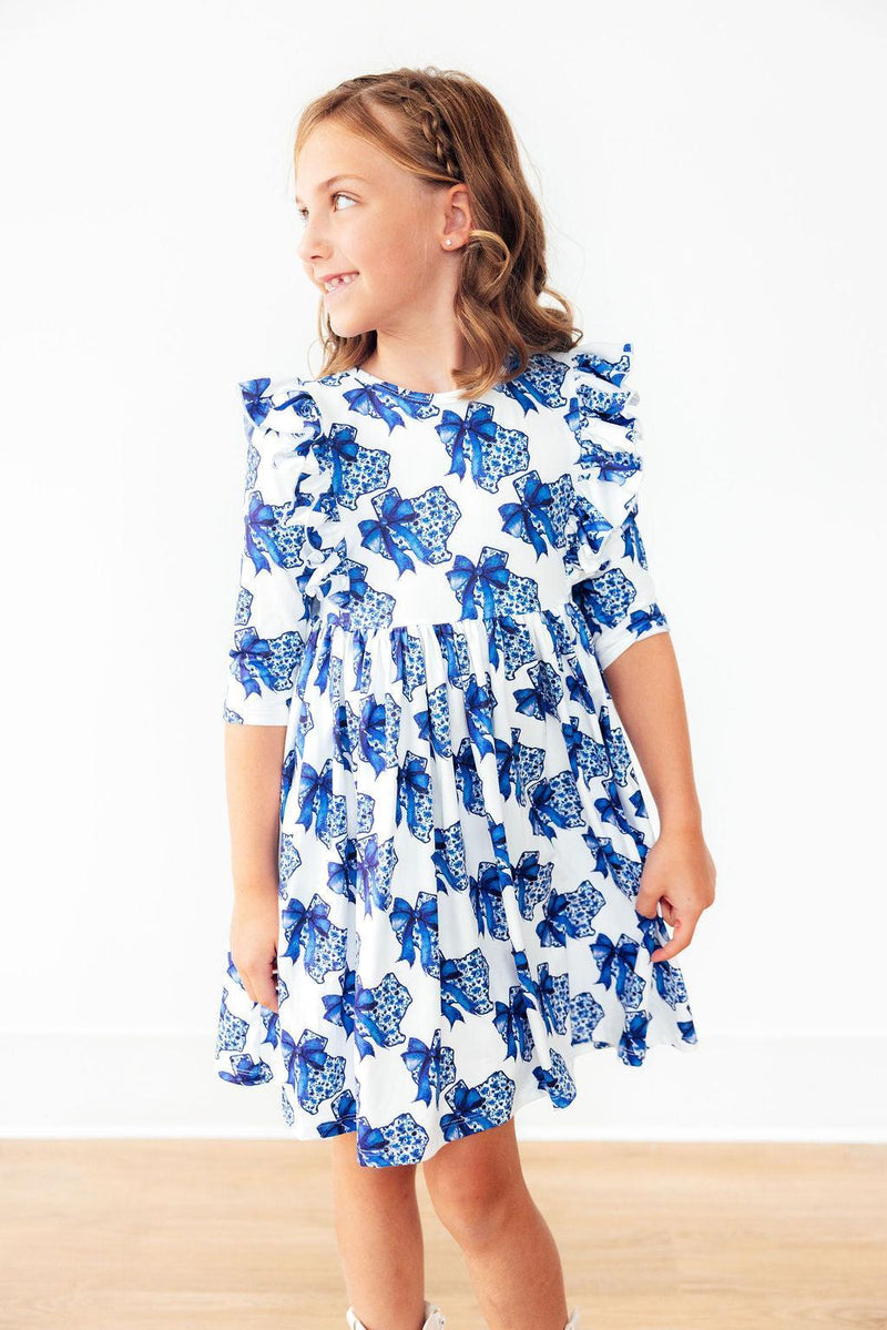 Side view of a smiling girl in the Lone Star 3/4 Sleeve Ruffle Twirl Dress, featuring an eye-catching blue bow design and elegant scoop back neckline.