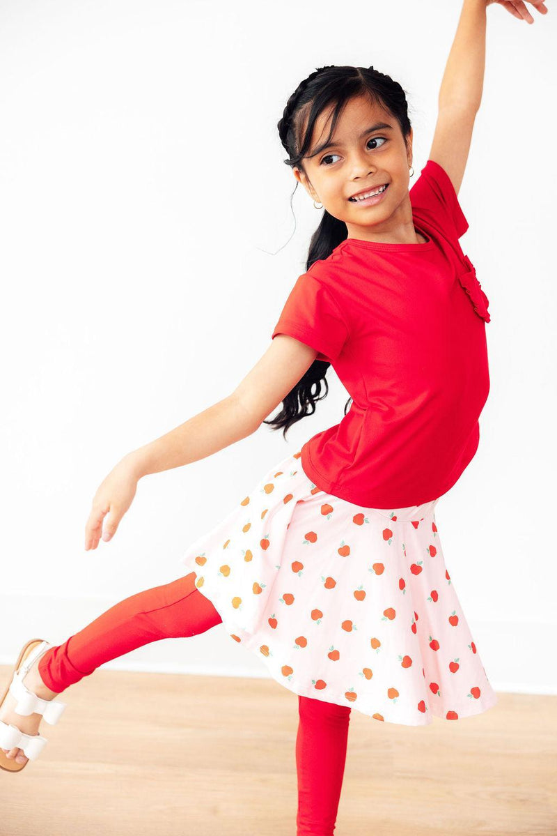 A joyful girl elegantly dancing in An Apple a Day Twirl Skeggings for Girls, featuring a playful skirt and leggings, illustrating the perfect choice in skeggings for girls.