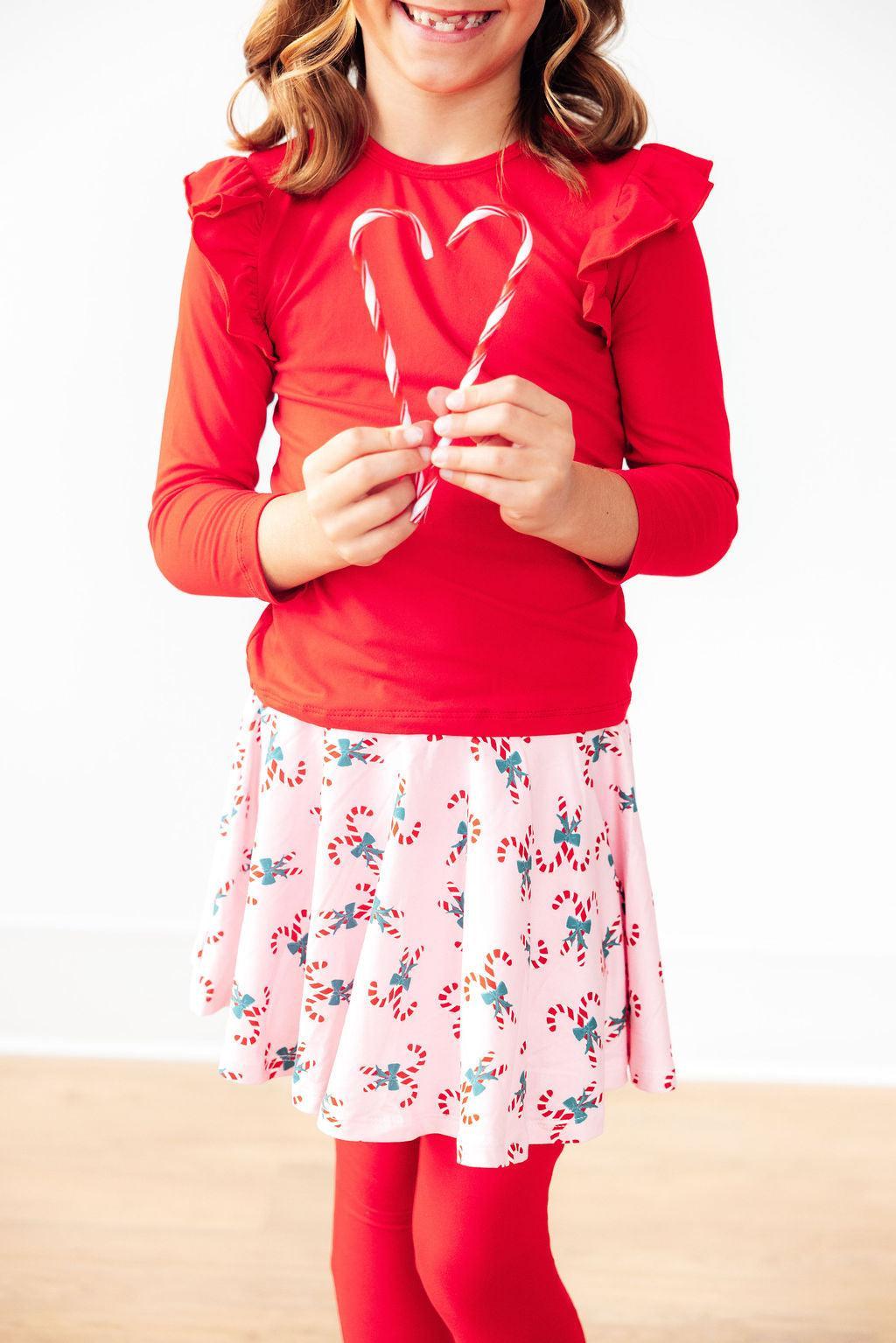 Girl wearing Candy Cane Cutie Twirl Skegging, smiling and holding candy canes, showcasing a fun red top with ruffled sleeves and a pink skirt adorned with candy designs.