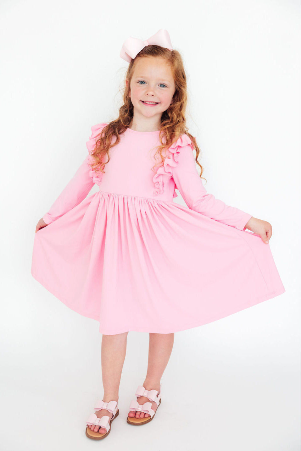 A cheerful young girl wearing a Bubblegum Pink L/S Ruffle Twirl Dress from Mila & Rose, playfully twirling with her hands raised. The dress features ruffles along the sleeves, in a soft bubblegum pink color, perfect for joyful occasions.