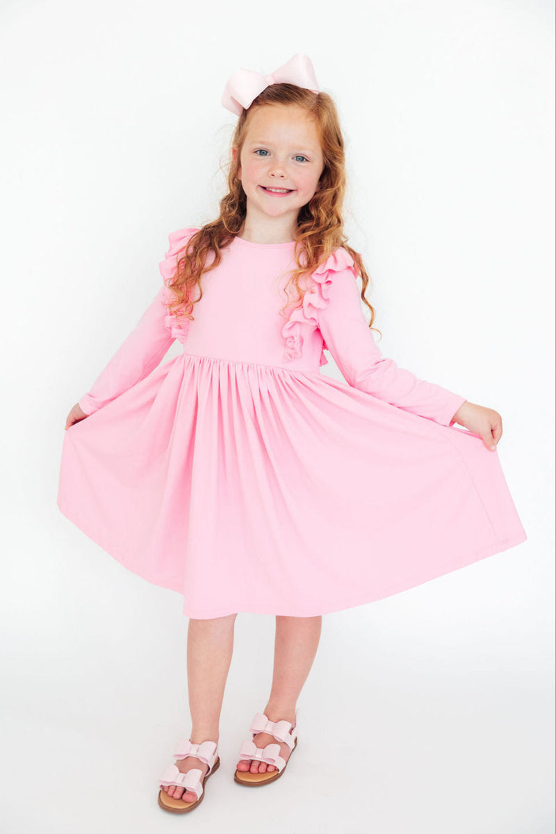 A cheerful young girl wearing a Bubblegum Pink L/S Ruffle Twirl Dress from Mila & Rose, playfully twirling with her hands raised. The dress features ruffles along the sleeves, in a soft bubblegum pink color, perfect for joyful occasions.