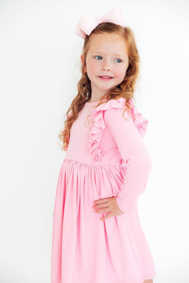 A smiling little girl wearing the Bubblegum Pink L/S Ruffle Twirl Dress, featuring ruffled sleeves and a twirling skirt, perfect for playdates and special occasions.