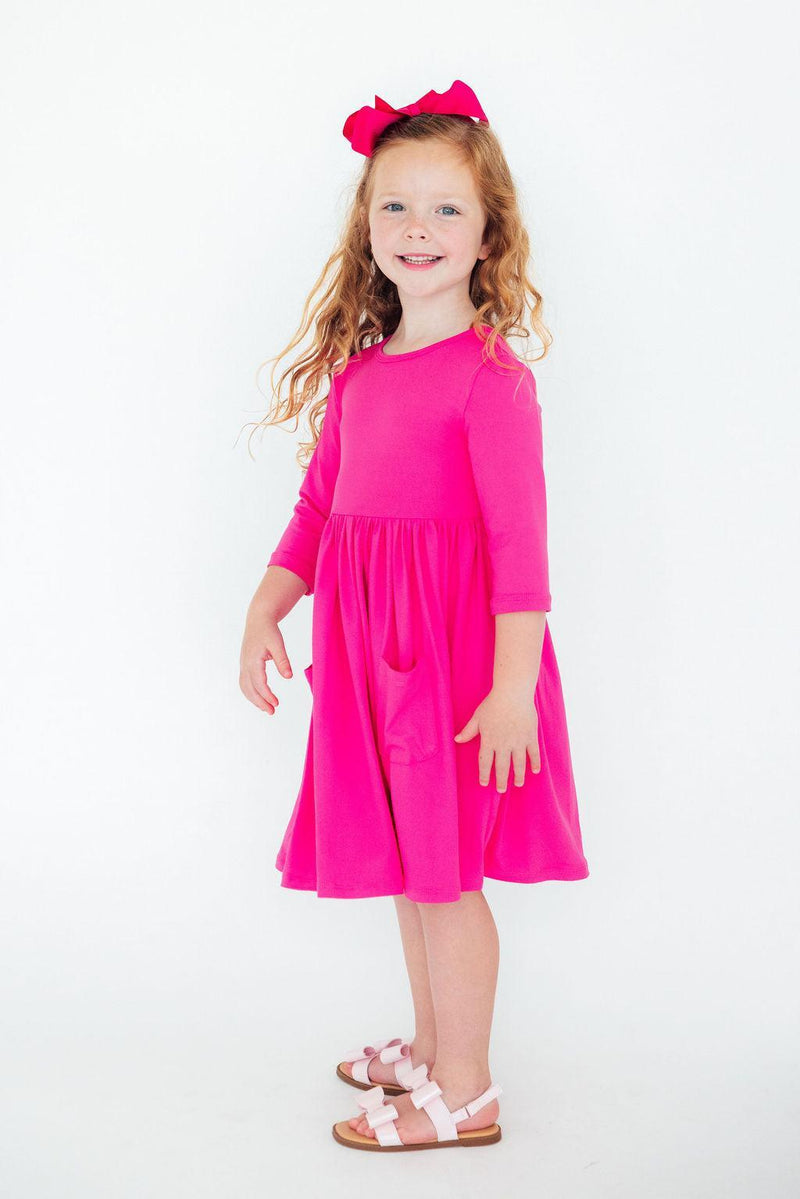 Smiling girl in a Hot Pink 3/4 Sleeve Pocket Twirl Dress, standing confidently with hands on hips, showcasing her fun fashion.