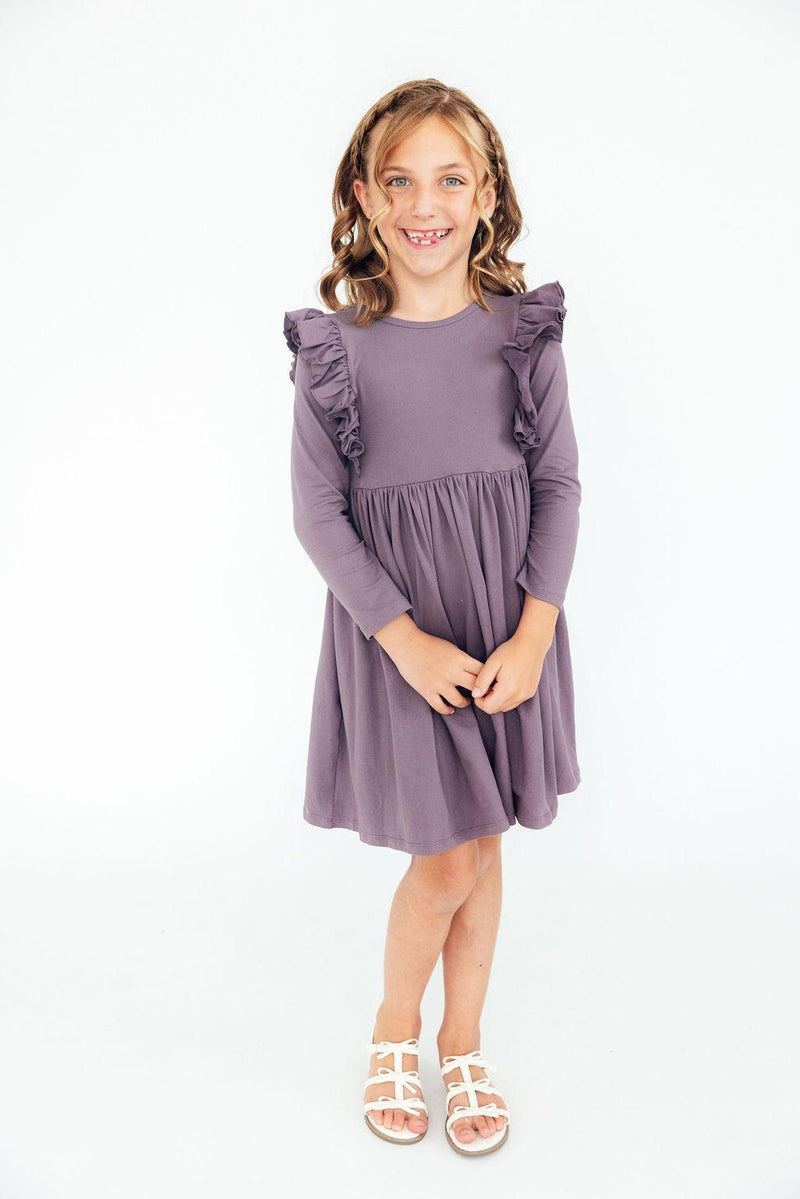 A smiling girl stands confidently in the Vintage Violet Long Sleeve Ruffle Twirl Dress, showcasing its playful ruffle design and comfortable fit.