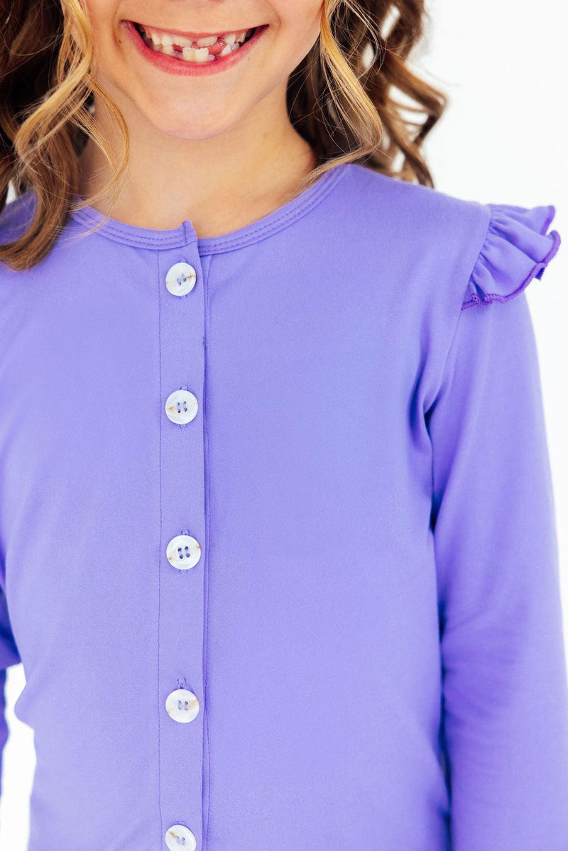 A cheerful girl wearing a Periwinkle Ruffle Cardigan, showcasing her bright smile and playful style with charming ruffle detailing and button accents.