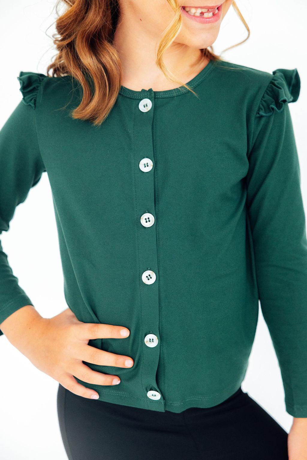 A young girl joyfully posing in a Spruce Ruffle Cardigan, showcasing the charming ruffle details and button front design. The cardigan's rich green color complements her casual outfit, making it perfect for playground adventures or family outings.