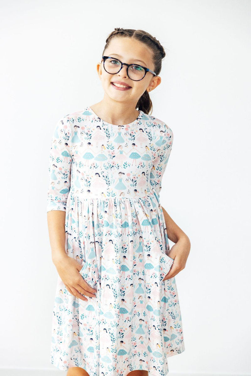 Cheerful girl posing in the Twinkle Toes 3/4 Sleeve Pocket Twirl Dress by Mila & Rose, showcasing a delightful design with vibrant dancers and flowers, ideal for fun and comfort.