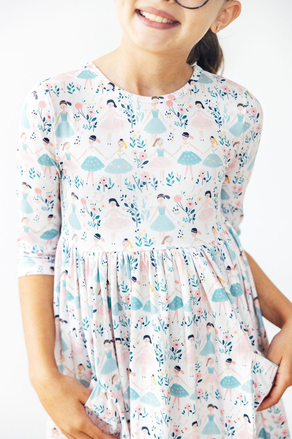 Young girl smiling while wearing the Twinkle Toes 3/4 Sleeve Pocket Twirl Dress from Mila & Rose, featuring a colorful print of dancers and floral patterns, perfect for playful twirling.