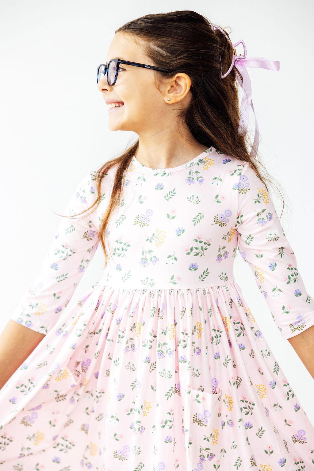 A cheerful girl wearing the Pastel Petals 3/4 Sleeve Pocket Twirl Dress, smiling and twirling, showcasing the playful floral design and her stylish glasses.