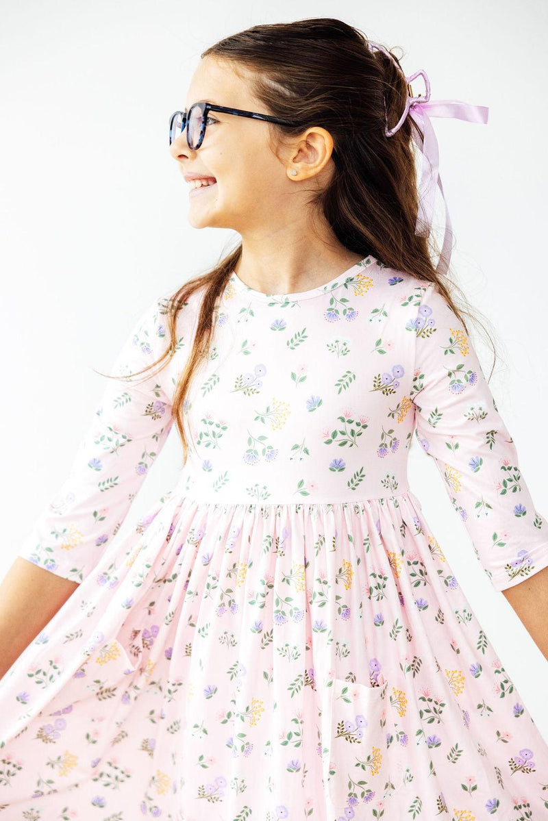A cheerful girl wearing the Pastel Petals 3/4 Sleeve Pocket Twirl Dress, smiling and twirling, showcasing the playful floral design and her stylish glasses.
