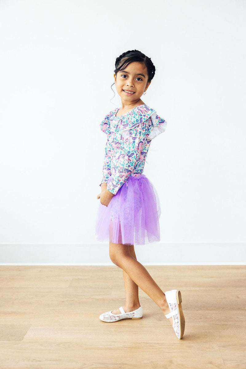 Side view of a young girl displaying Head in the Clouds Tutu Shorts, featuring a vibrant print and a whimsical purple tutu, looking playful and stylish.