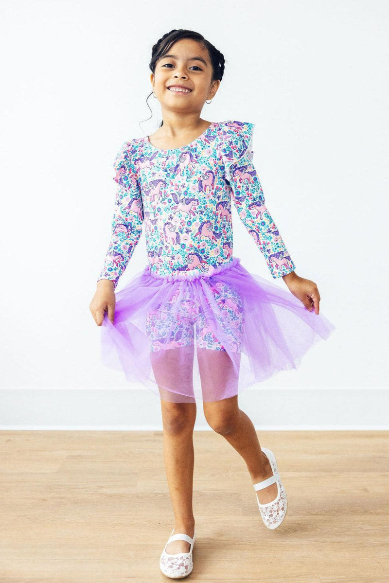 Head in the Clouds Tutu Shorts worn by a smiling young girl in a colorful dance outfit with a flouncy purple tutu overlay, striking a joyful pose.