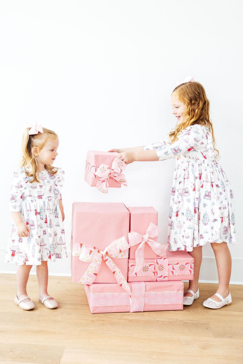 Two girls in Clara's Christmas 3/4 Sleeve Ruffle Twirl Dresses from Mila & Rose® joyfully interacting while surrounded by pink gift boxes.