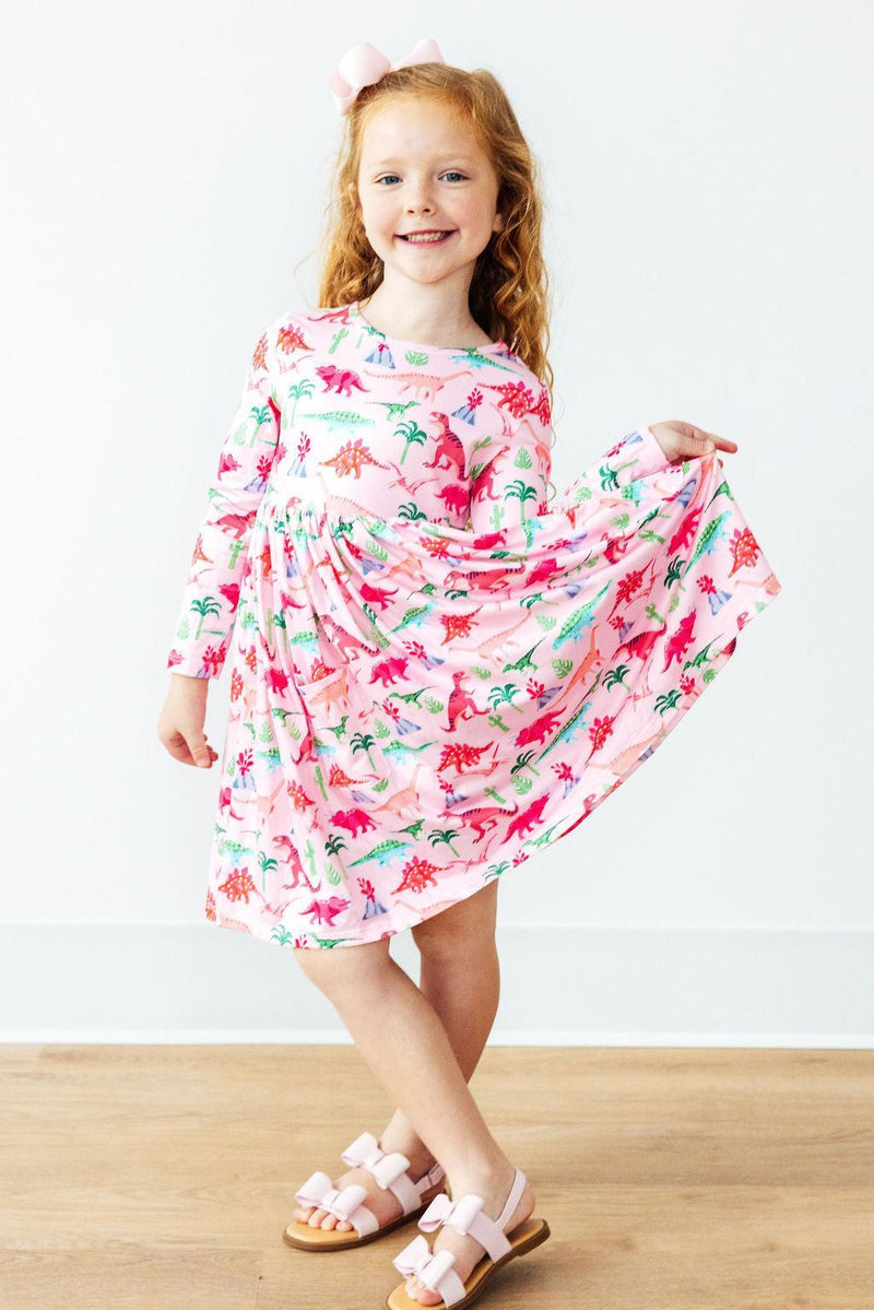 A cheerful child in the Pinkosaurus Rex Long Sleeve Pocket Twirl Dress by Mila & Rose®, twirling her skirt that features colorful dinosaurs, capturing the fun and playful spirit of the dress.
