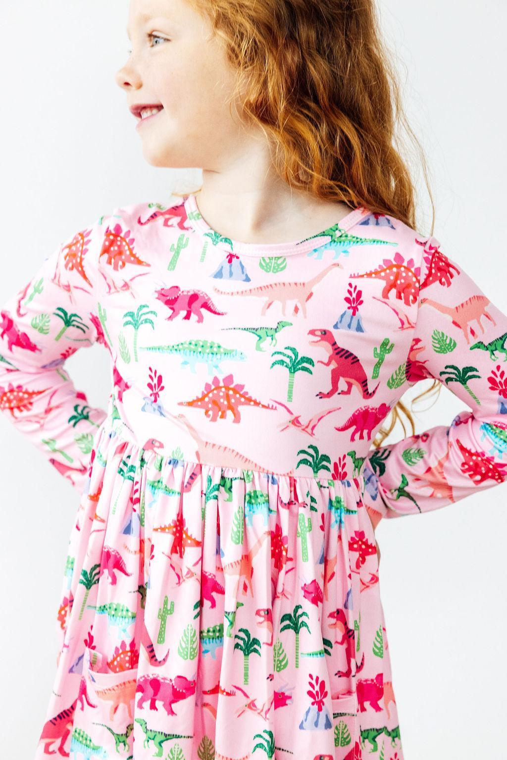 A young girl wearing the Pinkosaurus Rex Long Sleeve Pocket Twirl Dress by Mila & Rose®, showcasing a vibrant pink dinosaur print, smiling and posing confidently.