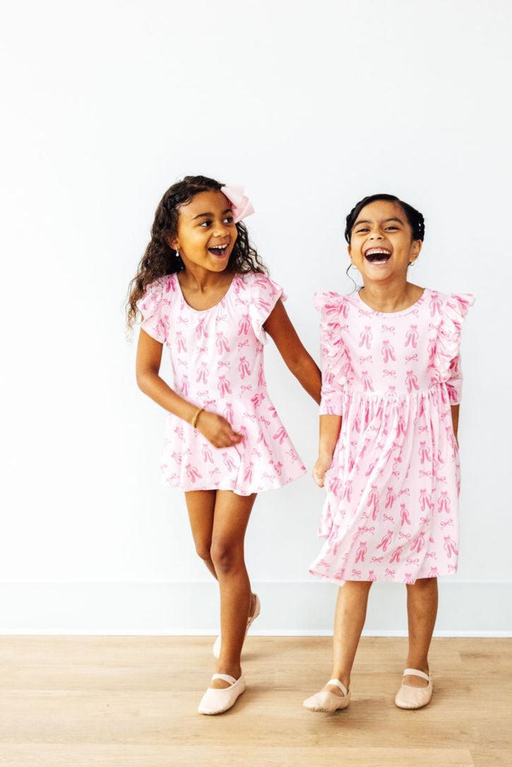 Two joyful girls in matching outfits featuring the Twirling en Pointe 3/4 Sleeve Ruffle Twirl Dress and a shorter version, radiating happiness while dressed in pink ballet-themed attire.