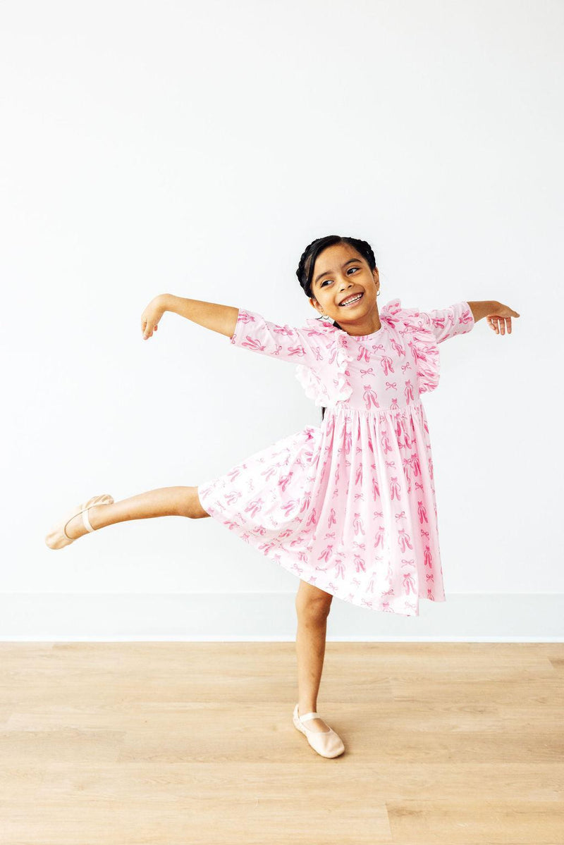 A smiling girl gracefully posing in the Twirling en Pointe 3/4 Sleeve Ruffle Twirl Dress, displaying the skirt's twirl ability and cute ballet slipper prints.