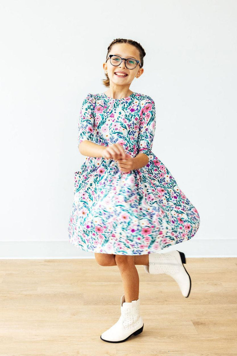 A joyful girl in a Vivid Garden 3/4 Sleeve Pocket Twirl Dress from Mila & Rose, spinning happily. The dress features a colorful floral pattern and a full twirl skirt, perfect for play and comfort.