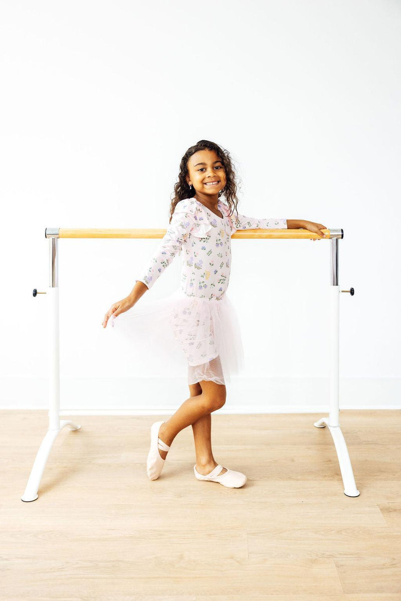 Smiling child in the Pastel Petals Long Sleeve Flutter Leotard standing by a ballet barre, demonstrating the versatility and charm of this dancewear.