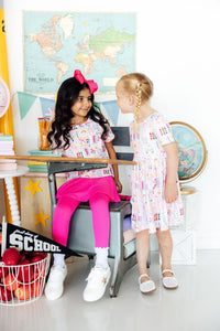 Two girls wear Art Class Short Sleeve Pocket Twirl Dresses, twirling in a classroom.