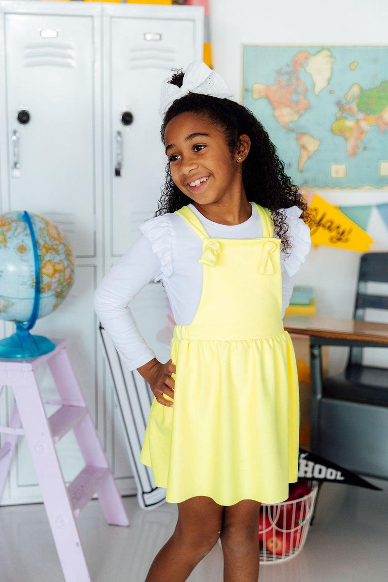 Girl wearing bright yellow faux suede pinafore with bow straps over a white top in a classroom.