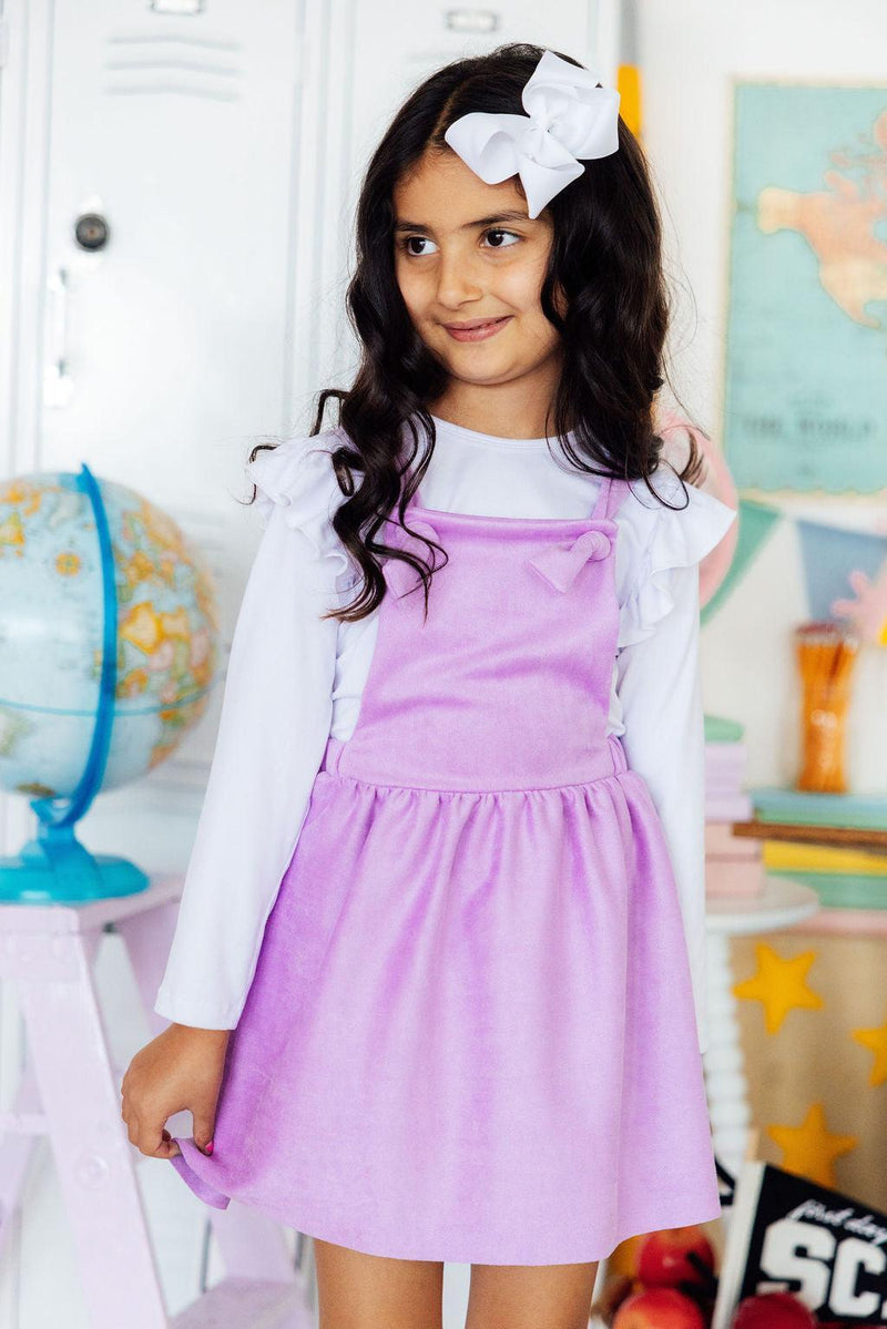 Girl wearing lavender faux suede pinafore over white top, with a large white bow in her hair.