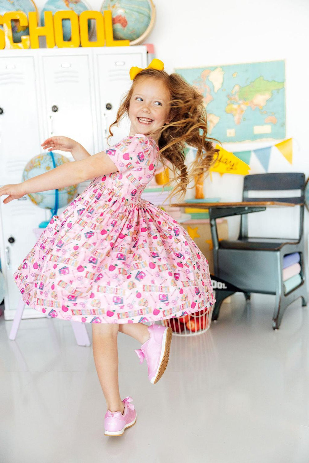Girl twirling in a pink Kindergarten Short Sleeve Pocket Twirl Dress in a bright classroom with lockers.